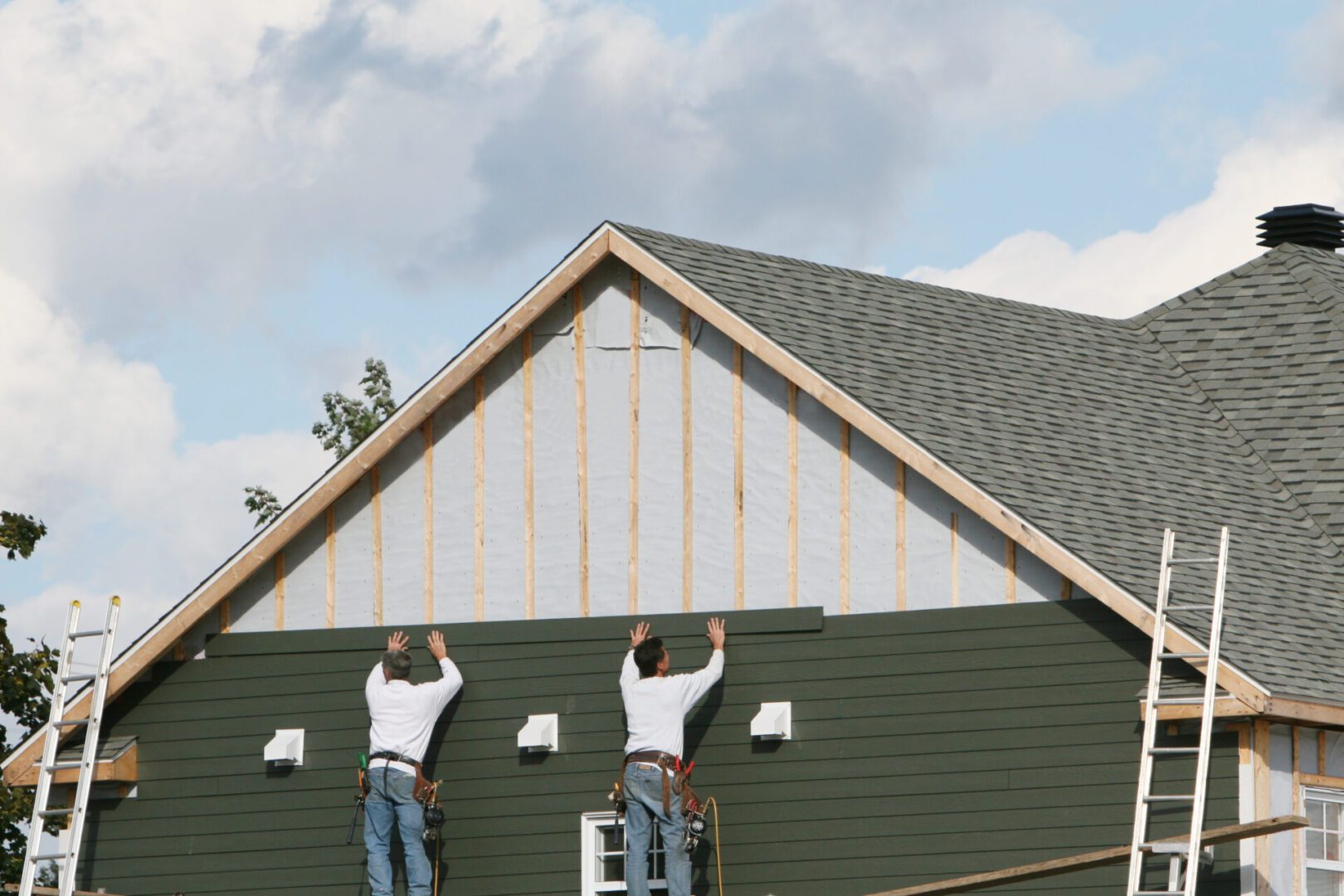 Two workers installing siding on a house's upper wall under a cloudy sky.